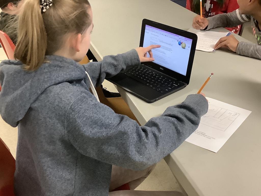 Young girl sitting at a table while pointing to her computer screen on her left side while writing on a piece of paper with her right hand.