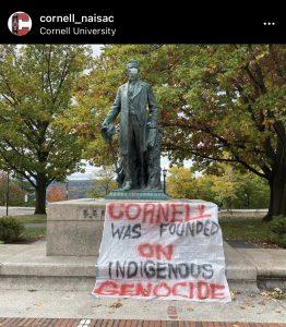 A banner stating "Cornell was founded on Indigenous genocide" hangs on a statue of Ezra Cornell