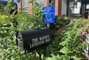 Black mailbox with the words "The Mayor's Listening Post" printed on the side. A blue sign is directly above the mailbox, in white letter it says, "Reimagining Public Safety in Ithaca and Tompkins County. Drop off input here or call 607 274 5465 or visiting bit.ly/2Jbfce9. Both the sign and the mailbox are surrounded by green shrubbery and some orange and yellow flowers. The leaves are partially covering the left side of the sign. 