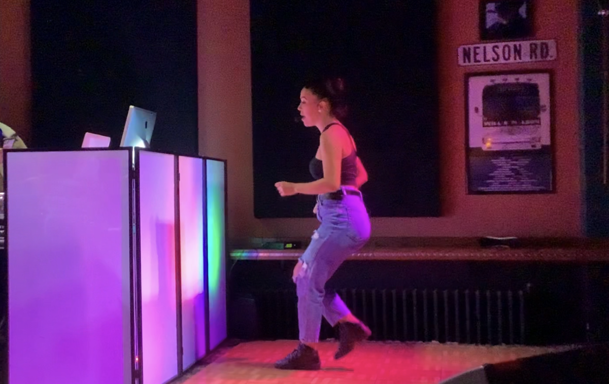 A woman in a black shirt and jeans teaches salsa steps on a stage in front of a colorful DJ booth