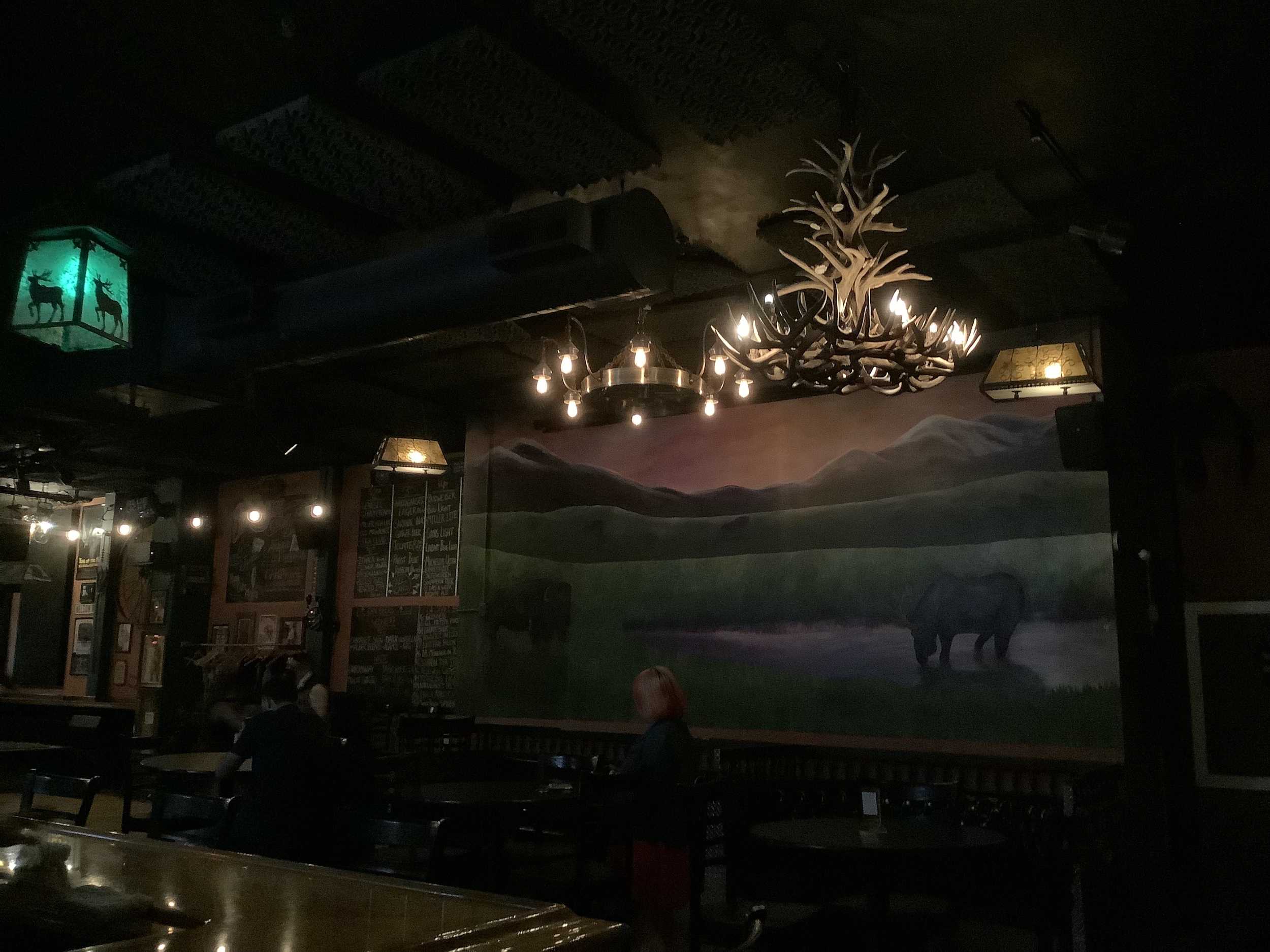 a prairie mural painted on the wall is lit up by the light from a chandelier made from antlers and light fixtures decorated with deer and elk
