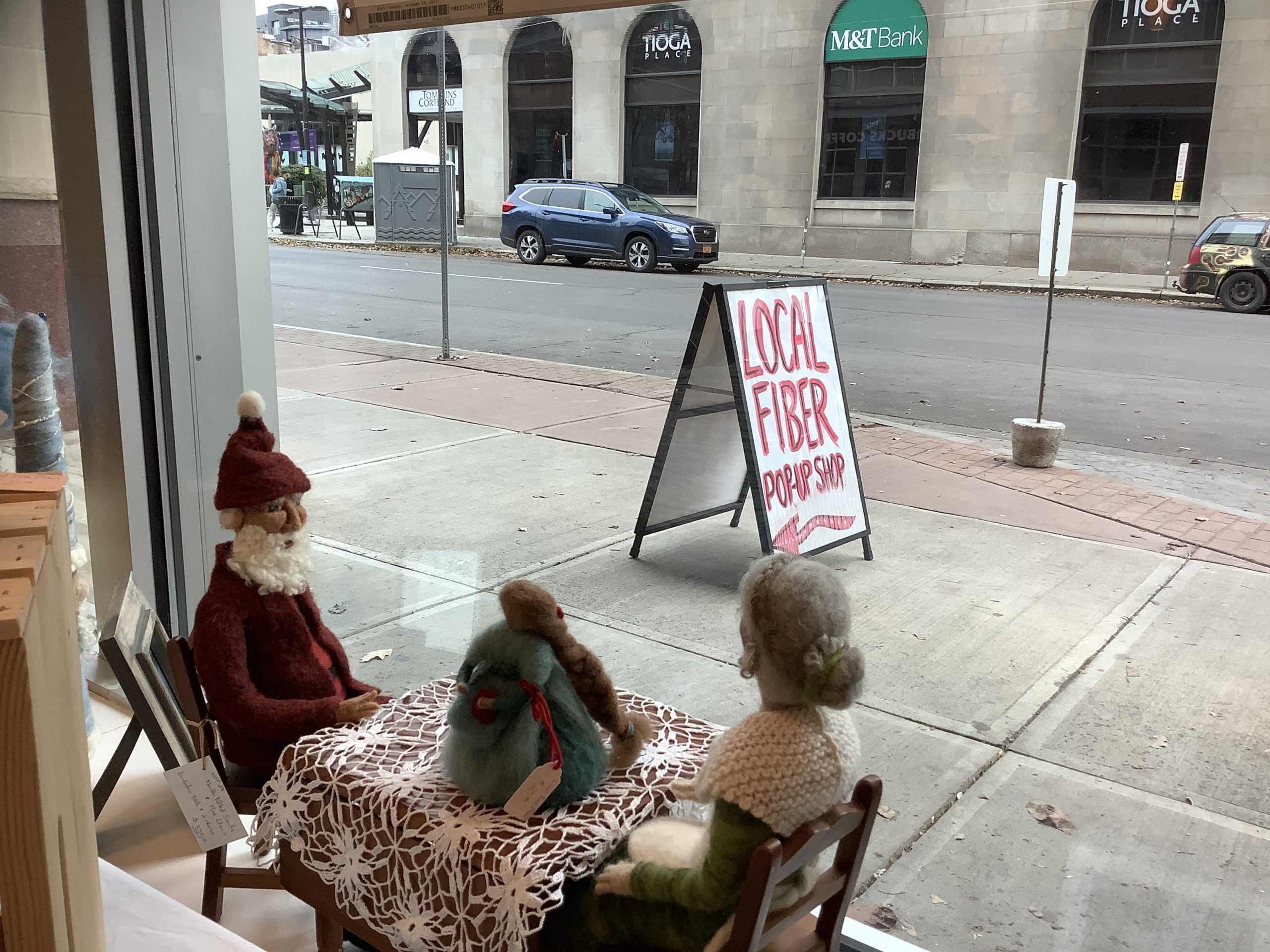 Picture of two wool dolls sitting in the window display from the interior of the store. The popup shop sign can be seen outside the window. 