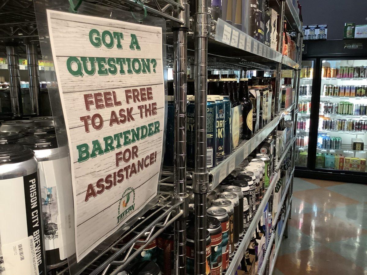 Shelves of craft beers with hundreds of different flavors and a sign for customers who may need assistance. Photo by Connor Ahern/Ithaca Week