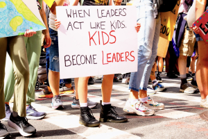 A young person holds a sign at a protest reading "when leaders act like kids, kids become leaders"