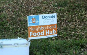 A sign in a grass field reads "Neighborhood Food Hub" and has the Friendship donations network logo.