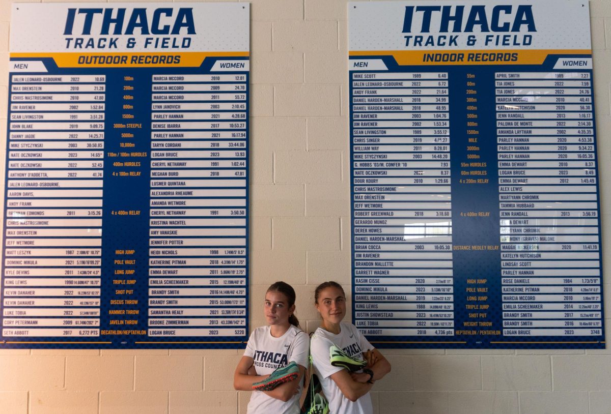 Freshman Raya Mount and Junior Lola Gitlin in Ithaca College cross country gear and racing spikes in front of the Ithaca College outdoor and indoor school record boards. Ithaca, New York. September 2025. (Hana Weisinger/Ithaca Week)